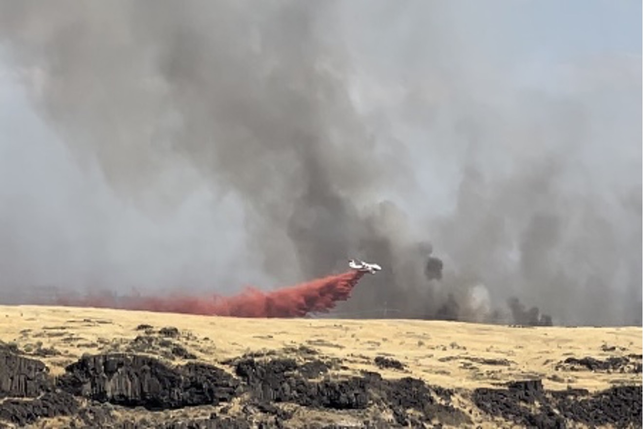 A jet dropping fire retardant across the field with black smoke in the background